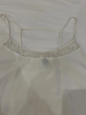 Vit linnetopp med spets från H&M - Säljer en vit linnetopp från H&M i storlek 36. Toppen har tunna axelband och en snygg spetsdetalj längs halsringningen. Materialet är lätt och luftigt, perfekt för varma dagar. Enkel och stilren design som är lätt att matcha med olika outfits.