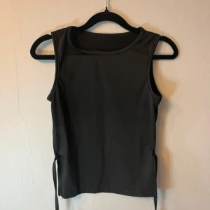 Svart ärmlös linnetopp med snörning - Svart ärmlös linnetopp med cool snörning i sidorna och öppen rygg. Toppen har en rak, något loose passform och är tillverkad i mjukt syntetmaterial. Perfekt för dig som gillar minimalistisk stil med en twist. Strl S 