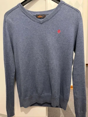 Blå v-ringad tröja Morris S - Blå v-ringad tröja från Morris i storlek S, tillverkad i 100% pima bomull. Tröjan har ett litet rött broderat emblem på bröstet och ribbade muddar vid hals, ärmslut och nederkant. Perfekt för dig som gillar klassisk och stilren look.