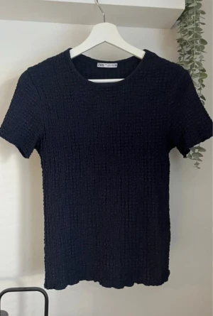Svart strukturerad topp från Zara - En svart topp från Zara med korta ärmar och rund halsringning. Toppen har en strukturerad, nästan krinklad yta som ger en cool och modern känsla. Perfekt att styla med jeans eller kjol för en enkel men trendig look.
