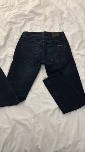 Mörkblå jeans från Nudie Jeans Co - Snygga mörkblå jeans från Nudie Jeans Co med klassisk femficksdesign och råa, avklippta benslut för en trendig look. Jeansen har subtila slitningar och Nudie-logga på bakfickan. Perfekta för dig som gillar en stilren men edgy vibe.