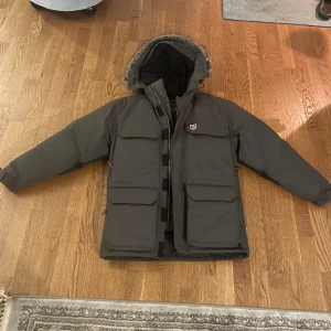 Mörkgrå dunjacka från Fjällräven - Säljer en mörkgrå dunjacka från Fjällräven med stor huva och snygg fuskpäls runt kanten. Jackan har flera stora fickorna framtill, dragkedja och knäppning, samt quiltad insida. Perfekt för kalla vinterdagar och har en riktigt schysst streetstil. Det är en XXS men väldigt stor i storlek så mer som en S!