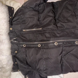 Svart Moose Knuckles dunjacka med päls - Svart dunjacka från Moose Knuckles med vit fuskpäls på huvan, broderad logga och hockeymotiv på insidan. Jackan har dragkedja, knappar och flera fickor, samt metall-detaljer på ärmen. Perfekt för kalla dagar och har en riktigt clean look.