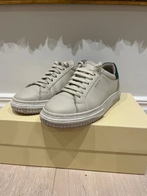 Vita sneakers från Axel Arigato - Snygga vita sneakers från Axel Arigato i skinn med klassisk rund tå och platt sula. Skorna har snörning och en grön detalj på hälen som ger en fräsch kontrast. Perfekta för dig som gillar stilrena och trendiga sneakers.