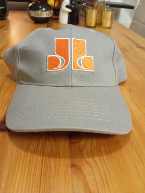 Grå keps med orange JL-logga - Snygg grå keps med stor orange JL-logga broderad framtill. Klassisk böjd skärm och justerbar baktill. Perfekt för dig som gillar stilrena accessoarer med en färgklick. Materialet är slitstark bomull som håller formen.