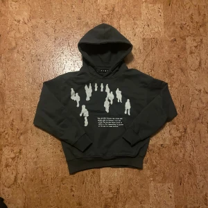 Svart hoodie från Admor XS - Svart hoodie från Admor i storlek XS, tillverkad i 100% bomull. Framsidan har ett coolt vitt tryck med silhuetter av människor och text. Tröjan har huva, ribbade muddar och klassisk loose passform. Perfekt för dig som gillar streetwear och grafiska detaljer.