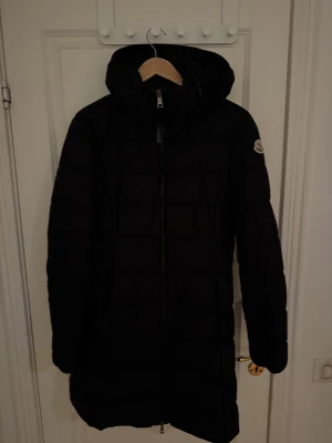 Svart lång dunjacka från Moncler - Svart lång dunjacka från Moncler med huva och quiltad design. Jackan har en snygg Moncler-logga på ärmen och dragkedja framtill. Perfekt för kalla dagar och har en stilren look som funkar till allt. Klassisk passform och riktigt varm.