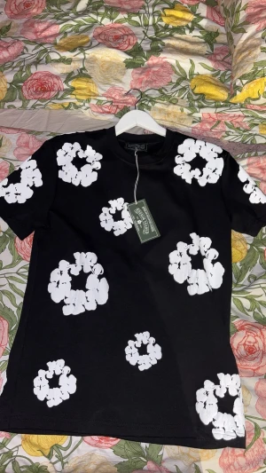 Svart t-shirt med vita blommor - Snygg svart t-shirt från Loyalty and Faith med coola vita blommönster över hela plagget. T-shirten har rund hals och korta ärmar, perfekt för dig som gillar streetstyle och vill sticka ut med ett unikt tryck.