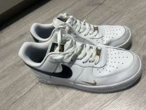 Nike Air Force 1 vit/guld sneakers - Nike Air Force 1 sneakers i vitt med svarta och guldfärgade detaljer. Klassisk låg modell med snörning, perforerad tå och platt sula. Swoosh-logga i guld på sidan och guldfärgad AIR-detalj på snörningen. Ovandel i skinn och textilfoder.