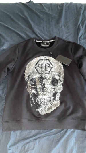 Svart Philipp Plein tröja med paljettdödskalle - Cool svart tröja från Philipp Plein med stor paljettdödskalle i silver på bröstet och logga upptill. Rund hals, långärmad och ribbade muddar. Snyggt statement-plagg med exklusiv vibe och detaljerad design.