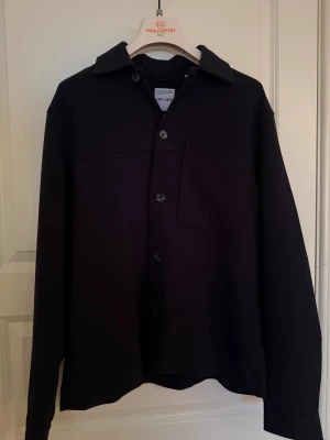 Svart overshirt från Only & Sons - Stilren svart overshirt från Only & Sons med klassisk krage, bröstficka och knäppning framtill. Perfekt för lager-på-lager och enkel att matcha med det mesta. Långärmad och i mjukt material för en clean look. Sitter mer som en stor small/ liten medium