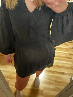 Svart kort klänning från Nelly - Supersöt svart kort klänning från Nelly med långa puffärmar, volangdetaljer och v-ringning. Klänningen har ett lätt transparent tyg med diskret struktur och markerad midja. Perfekt för dig som gillar en feminin och trendig look.