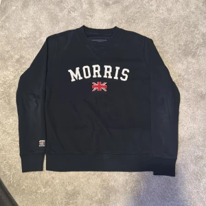 Mörkblå sweatshirt från Morris XS/S - Snygg mörkblå sweatshirt från Morris i storlek XS. Tröjan har vit broderad text och en röd/vit Union Jack-flagga framtill. Rund hals, ribbade muddar och en liten patch på ärmen. Perfekt för dig som gillar klassisk brittisk stil.