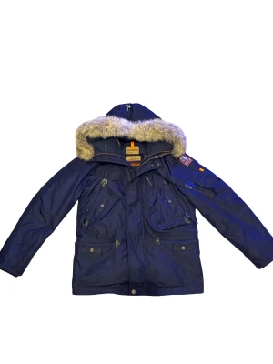 Parajumpers Anchorage mörkblå parkas - Säljer en mörkblå Parajumpers Anchorage parkas med avtagbar päls på huvan, flera fickor med dragkedja och knappar, samt ikonisk PJS-patch på ärmen. Jackan har lång ärm, gul rem vid halsen och robust design som håller dig varm hela vintern. Retail: 11799kr köpt från care of carl