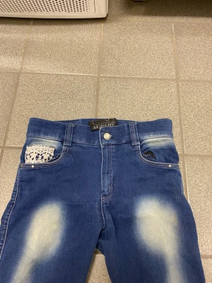 Blå ridbyxor från Horselife med spets - Snygga blå ridbyxor från Horselife med ljusa slitningar och dekorativa spetsdetaljer på fickorna fram och bak. Byxorna har smal passform, förstärkta partier vid bensluten och coola utskärningar längs benen. Perfekta för dig som gillar unik stil i stallet.