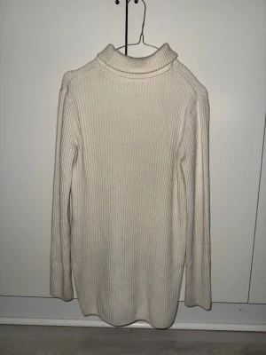 Beige ribbad polotröja - Säljer en beige ribbad polotröja med lång ärm och hög krage och snitt på ena sidan. Tröjan är stickad och har en mjuk, skön känsla som passar perfekt när det är kyligt ute. Enkel och stilren design som är lätt att matcha med olika outfits.