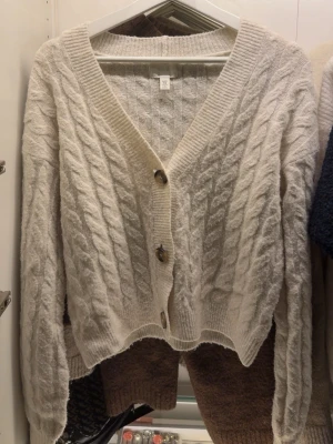 Vit kabelstickad kofta från H&M - Mysig vit kabelstickad kofta från H&M i storlek XS. Koftan har v-ringning, stora knappar framtill och ribbade muddar. Perfekt för lager-på-lager och chill dagar. Snyggt mönster och skön passform.