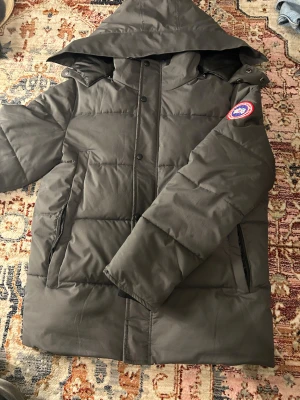 Mörkgrå dunjacka Canada Goose L - Säljer en mörkgrå dunjacka från Canada Goose i storlek L. Jackan har huva, dragkedja och knappar framtill samt två sidofickor. Klassisk Canada Goose-märke på ärmen. Perfekt för kalla dagar och riktigt snygg stil.