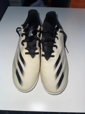 Adidas inneskor storlek 42 beige och svart - Adidas inneskor i storlek 42 med beige och svart färg. Skorna har snörning och en yttersula designad för inomhusbruk.