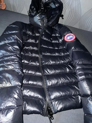 Svart dunjacka från Canada Goose - Säljer en svart dunjacka från Canada Goose med klassisk logga på ärmen. Jackan har quiltad design, huva och hel dragkedja framtill. Materialet är glansigt och känns lätt men ändå varmt, perfekt för kalla dagar. Passformen är normal och jackan har långa ärmar. Pris ej hugget i sten