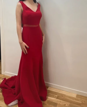 Röd långklänning med meshdetalj - Säljer en elegant röd långklänning med v-ringning och ärmlös design. Klänningen har en snygg meshdetalj i midjan och ett långt, svepande släp. Perfekt för bal eller festliga tillfällen. Figurnära passform som framhäver siluetten.