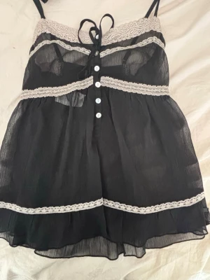 Svart och vit spetslinnetopp med knyt - Säljer en svart linnetopp med tunna axelband och transparent tyg. Toppen har vita spetsdetaljer längs bröstet, midjan och nederkanten, samt knyt framtill och dekorativa knappar. Perfekt för dig som gillar romantisk och feminin stil.