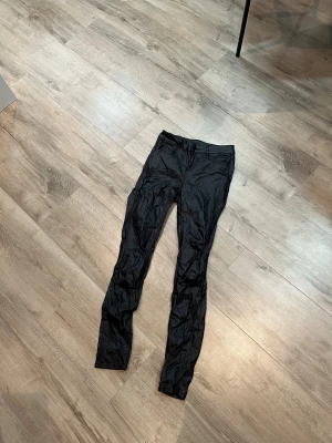 Svarta jeans byxor från VILA - Snygga svarta byxor från VILA i skinny modell. De har en glansig finish och är tillverkade i ett syntetmaterial som ger en cool look. Byxorna har klassisk midja och smal passform, perfekta för dig som gillar en stilren och modern vibe.