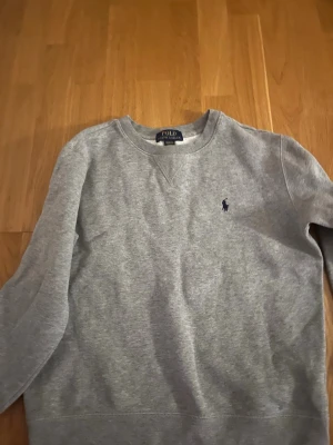 Grå sweatshirt från Polo Ralph Lauren - Snygg grå sweatshirt från Polo Ralph Lauren med klassisk rund hals och det ikoniska lilla broderade logot på bröstet. Tröjan har långärmad design och är gjord i mjuk bomull, perfekt för chill dagar. Enkel och stilren look som funkar till jeans eller joggers.