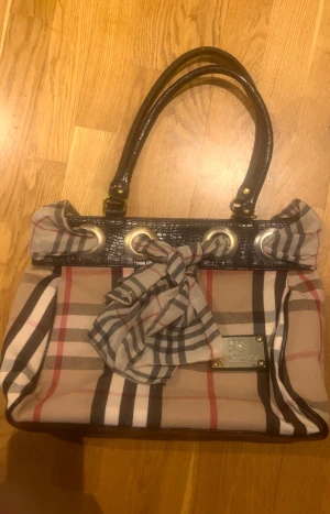 Rutig handväska från Burberry - Snygg handväska från Burberry med klassiskt rutigt mönster i beige, svart, vitt och rött. Väskan har svarta handtag i skinn, stora guldfärgade metalldetaljer och en dekorativ rosett framtill. Perfekt för dig som vill ha en ikonisk accessoar.