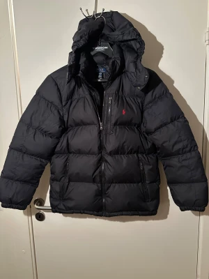Svart pufferjacka Polo Ralph Lauren XL - Svart pufferjacka från Polo Ralph Lauren i storlek XL barn. Jackan har huva, dragkedja framtill och två sidofickor med dragkedja. Klassisk röd Polo-logga på bröstet. Perfekt för kalla dagar med sin tjocka, quiltade design. Skriv vid minsta funderingar😁
