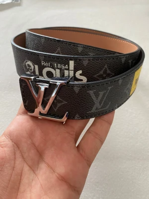 Iv monogram bälte - Snyggt Iv bälte med klassiskt monogrammönster i mörkbrunt och grått, detaljer i gult och blått samt stor Iv-spänne i silver. Bältet är gjort i skinn med insida i beige och har coola tryck och loggor längs bandet. Perfekt accessoar för att lyfta din outfit.