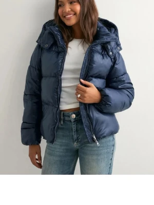 Mörkblå pufferjacka med huva - Snygg mörkblå pufferjacka med stor huva och dragkedja framtill. Jackan har quiltad design och är tillverkad i glansig syntet, perfekt för kalla dagar. Modellen är normal i passningen och har långa ärmar. Enkel och stilren look som funkar till jeans och t-shirt.