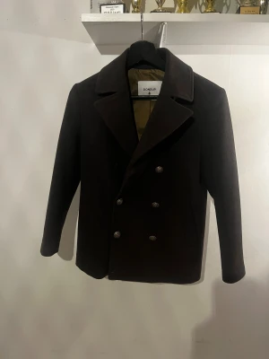 Skepparkavaj från Dondup - Stilren mörkbrun skepparkavaj från Dondup med klassisk dubbelknäppning och snygga metallknappar med stjärndetalj. Jackan har bred krage och är helfodrad med olivgrönt innerfoder. Perfekt för dig som vill ha en tidlös och clean look.