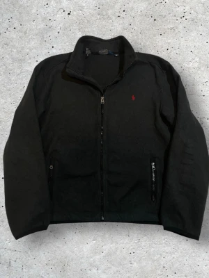 Svart fleecekofta från Polo Ralph Lauren XL  - Svart fleecekofta från Polo Ralph Lauren i storlek XL. Tröjan har hel dragkedja framtill, ståkrage och två fickor med dragkedja. Klassisk röd Polo-logga broderad på bröstet. Något mindre i storleken 