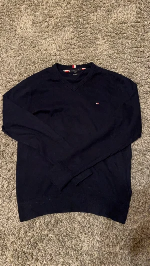 Mörkblå tröja från Tommy Hilfiger - Mörkblå långärmad tröja från Tommy Hilfiger i en mjuk mix av pima bomull och kashmir. Klassisk v-ringning och liten broderad logga på bröstet. Perfekt för dig som gillar stilrena och bekväma plagg.