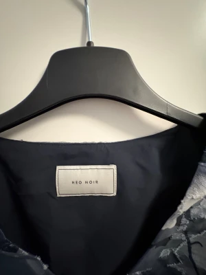 Blommig blus med puffärm från Neo Noir - Säljer en snygg blus från Neo Noir med blommigt mönster i mörkblått, grått och vitt. Blusen har v-ringning, korta puffärmar och en slits i sidan. Materialet är lätt och har en lite glansig yta som känns lyxig. Perfekt för dig som gillar statement-ärmar och coola prints.