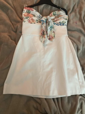 Vit kort klänning med blommig rosett Zara - Supersöt vit kort klänning från Zara med blommigt mönster upptill och en stor rosett framtill. Axelbandslös design och lätt A-linjeformad kjol. Perfekt för sommarens alla tillfällen, med fräscha färger och mjukt material.