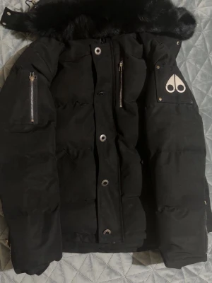 Svart Moose Knuckles pufferjacka - Säljer en svart pufferjacka från Moose Knuckles med fet päls på huvan, silvriga dragkedjor och metallpatch på ärmen. Jackan har broderad insida med hockeymotiv och snygga detaljer som stora knappar och fickor. Perfekt för dig som vill ha en varm och stilren vinterjacka. Passar de som har storlek L då mosse knuckles är liten i storlek 