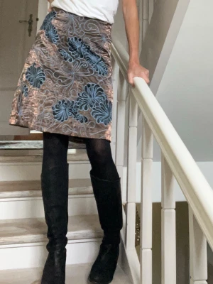 Kjol från French Connection - Supersnygg kjol från French Connection med bottenfärg i brun/koppar och asymmetriskt broderi i svart och ljusblått. Kjolen har en A-linjeformad siluett och är gjord i glansigt satinmaterial som ger en lyxig känsla. Osynlig dragkedja bak. Passar så bra till både svart, vitt/beige och bruna toner. Funkar alla årstider, ett riktigt kvalitetskap från ett klassiskt märke. Obetydligt använd! 