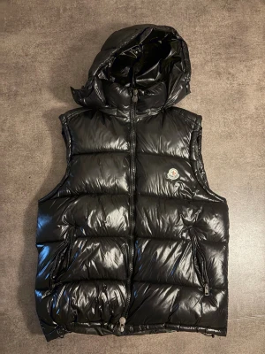 Svart dunväst från Moncler med huva - Svart, glansig dunväst från Moncler med avtagbar huva och två fickor med dragkedja. Västen har ett quiltat mönster och klassisk Moncler-logga på bröstet. Perfekt för lager-på-lager och streetstyle. Stängs med dragkedja framtill.