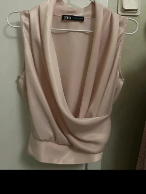  Ny Champagnefärgad omlottopp från Zara - Säljer en elegant champagnefärgad omlottopp från Zara. Toppen är ärmlös med draperad v-ringning och bred linning nedtill. Materialet har en satinliknande finish som ger en lyxig känsla. Perfekt för dig som gillar stilrena och trendiga plagg.