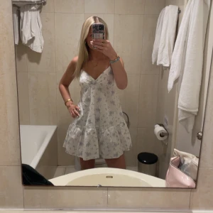 Vit blommig kortklänning med volang - Supersöt vit kortklänning med ljusblå blommigt mönster och volang nedtill. Klänningen har v-ringad hals, ärmlös design med tunna axelband och en luftig passform som ger en chill vibe. Perfekt för varma dagar och sommarhäng med vänner.