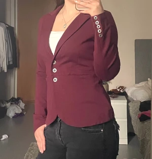 Vinröd kavaj från House of Lola - Snygg vinröd kavaj från House Of Lola med figurnära passform, klassisk krage och tre knappar framtill. Långa ärmar med dekorativa knappar vid ärmslut och broderad detalj av en älva i nacken. Mjukt collegetyg som är stretchigt och skönt att bära. Nypris ca: 900kr. Missfärgad vid insidan av kragen, men inget som syns vid användning.