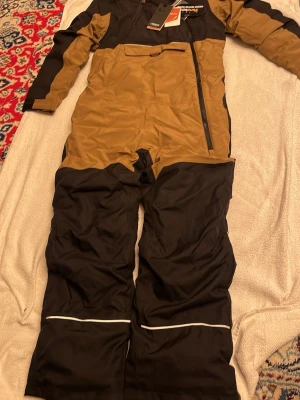 Bruna och svarta täckbyxor Helly Hansen - Säljer ett par täckbyxor från Helly Hansen i brunt och svart med vita reflexdetaljer vid bensluten. Byxorna har dragkedja på sidan och är tillverkade i slitstarkt syntetmaterial, perfekta för kalla dagar. Snygg design med paneler och praktisk ficka framtill.