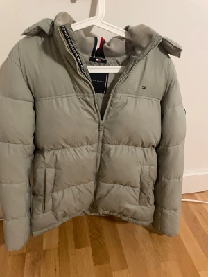 Ljusgrå pufferjacka Tommy Hilfiger - Säljer en ljusgrå pufferjacka från Tommy Hilfiger med huva. Perfekt för kalla dagar nu i vinter. Säljer den billigt då den tar för mycket plats i min garderob eftersom jag ej använder denna.                 Köpt på zalando för 1400kr.