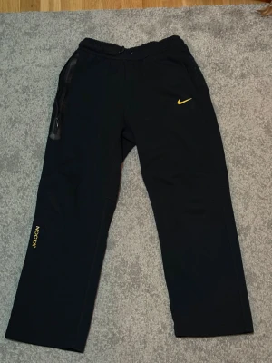 Nike byxor  - Svarta mjukisbyxor från Nike NOCTA i storlek S med gul logga och detaljer. Byxorna har elastisk midja med snörning (som ena hamnat utanför hålet), raka ben och praktiska fickor med dragkedja. Materialet är mjukt och passar perfekt för chill eller träning. Snygga NOCTA-tryck på benet och diskreta broderade detaljer.