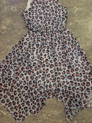 Leopardmönstrad kortklänning - Snygg kortklänning med leopardmönster i svart, beige och orange. Klänningen har ärmlös design, markerad midja och asymmetrisk nederkant som ger en cool och trendig look. Perfekt för dig som vill sticka ut med ett djurmönster.