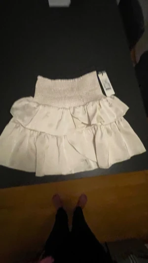 Vit volangkjol i satin från Neo Noir - Supersöt kort kjol från Neo Noir i pärlvit satin med bred smock i midjan och dubbla volanger. Kjolen har en glansig finish och känns riktigt lyxig och flowy. Perfekt för dig som vill ha en feminin och trendig look.