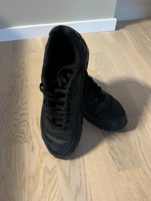Nike Air Max 90 helsvarta sneakers - Ordinarie pris 1900. Använda 2 gånger. Säljer ett par helsvarta Nike Air Max 90 sneakers med snygg skinnfinish och klassisk siluett. Skorna har platt sula, rund tå och synlig Air Max-dämpning i sulan. Diskret Nike-logga på sidan och snörning framtill. Perfekt för dig som gillar stilrena och sportiga sneakers.