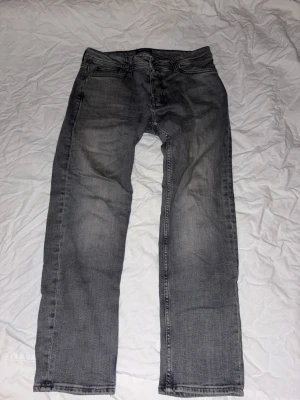 Gråa Jack&Jones jeans - Säljer ett par grå jeans från Jack&Jones med klassisk femficksdesign och raka ben. Jeansen har en snygg tvättad look och svarta detaljer på bakfickan. Passar dig som gillar stilrena och avslappnade jeans med straight fit.